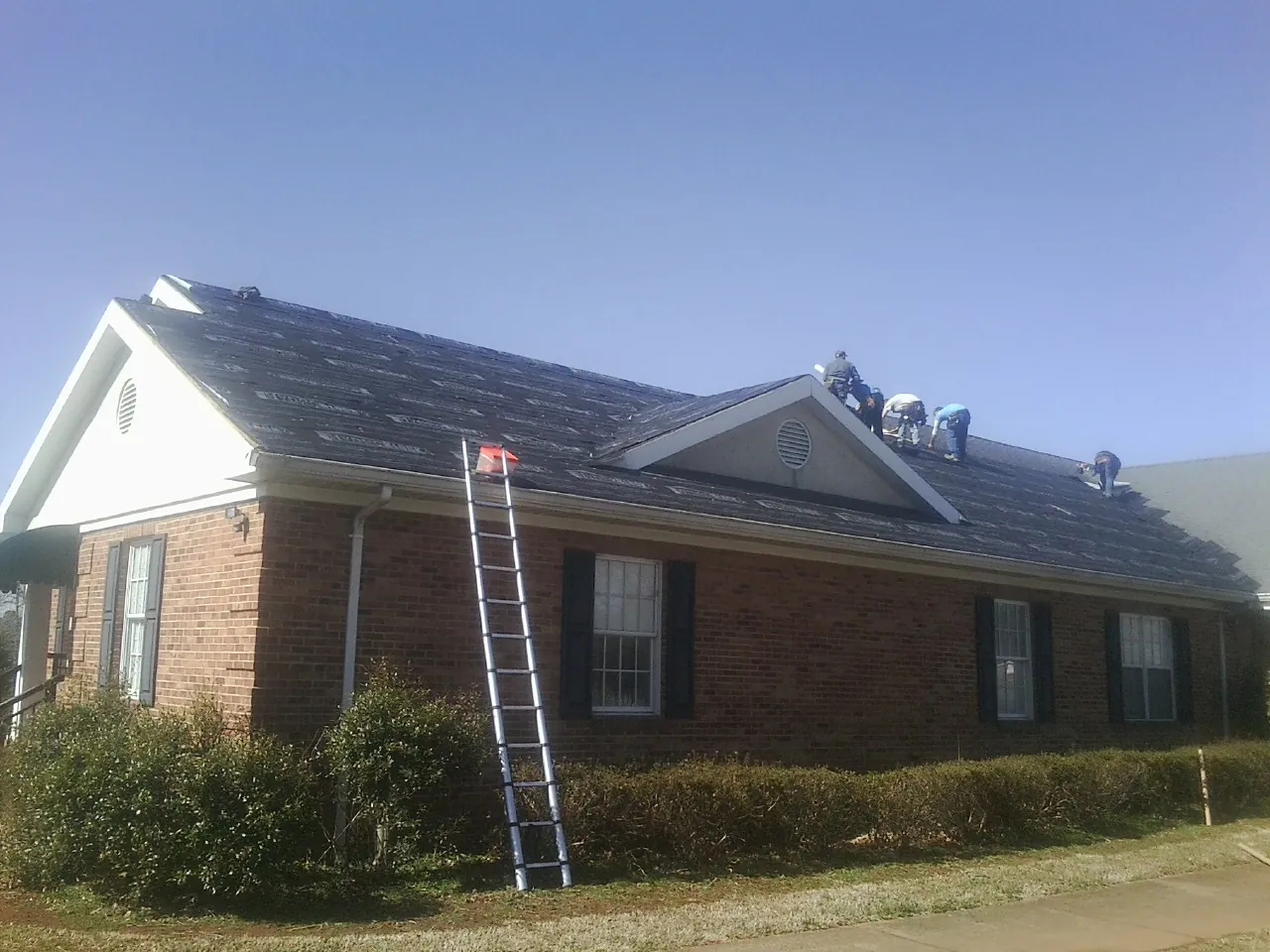 Crew tearing shingles off a brick ranch roof with a ladder set — Emergency Roof Repair work by our crew in Jemison