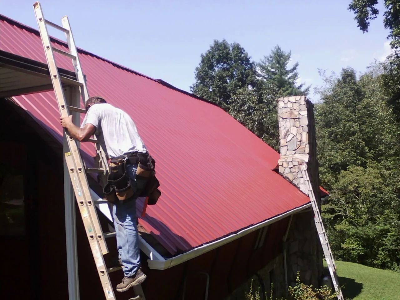 Roofer on a ladder beside a red metal roof and stone chimney — Soffit & Fascia Repair by Roofing Brothers in Jemison